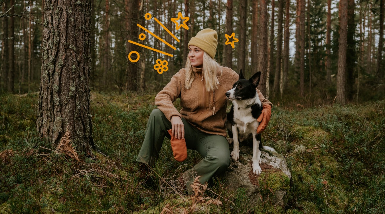 Frau mit blonden Haaren sitzt im Wald auf einem Stein mit ihrem Hund im Arm